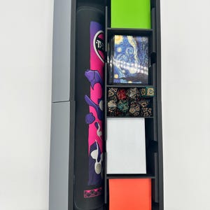 May include: A black and gray storage tower with multiple compartments. The tower contains a rolled-up playmat with pink and purple accents, green and orange card sleeves, and a deck of cards featuring Van Gogh's "Starry Night" painting. Also visible are dice.