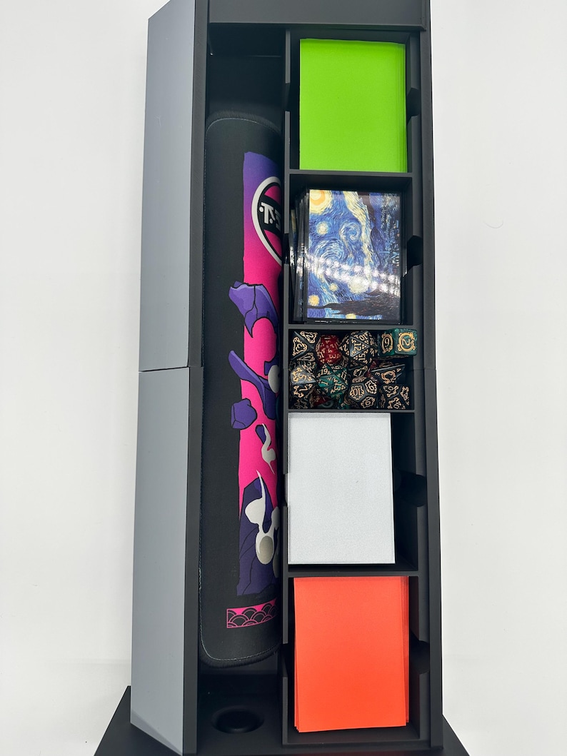May include: A black and gray storage tower with multiple compartments. The tower contains a rolled-up playmat with pink and purple accents, green and orange card sleeves, and a deck of cards featuring Van Gogh's "Starry Night" painting. Also visible are dice.