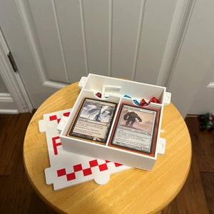 Op de afbeelding: Witte plastic doos met twee compartimenten voor het bewaren van speelkaarten en dobbelstenen. De doos staat op een wit-rood geruit oppervlak. De kaarten zijn Magic: The Gathering kaarten. De tekst op de kaarten luidt "Arctic Chasm" en "Stone Golem".