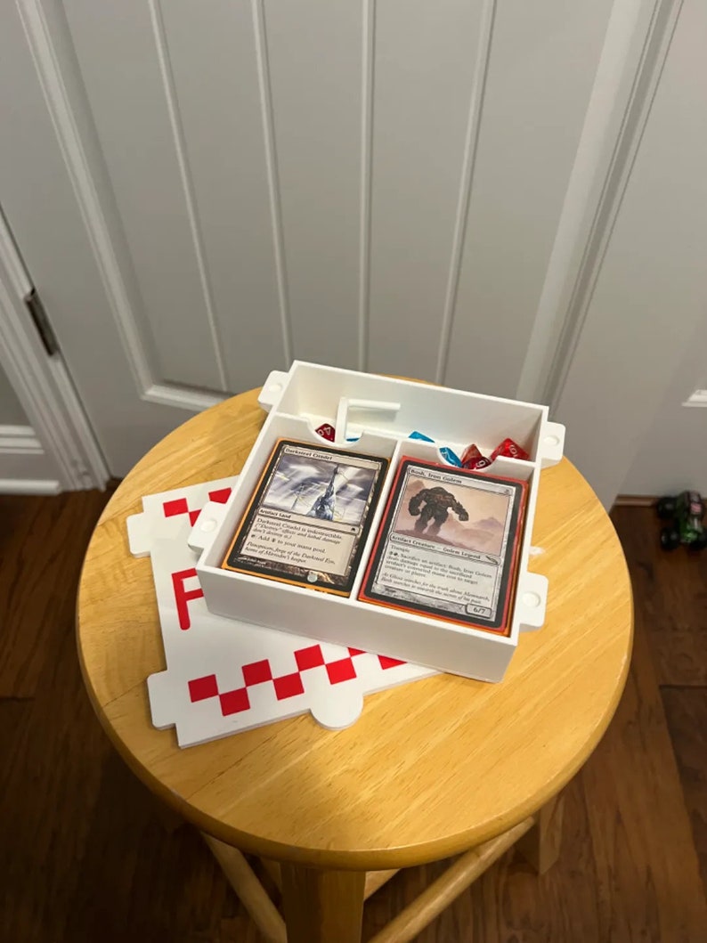 Op de afbeelding: Witte plastic doos met twee compartimenten voor het bewaren van speelkaarten en dobbelstenen. De doos staat op een wit-rood geruit oppervlak. De kaarten zijn Magic: The Gathering kaarten. De tekst op de kaarten luidt "Arctic Chasm" en "Stone Golem".