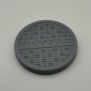 May include: A gray, circular coaster with a manhole cover design. The text "CITY SEWER" is embossed in the center. The coaster has a textured surface and a raised edge.