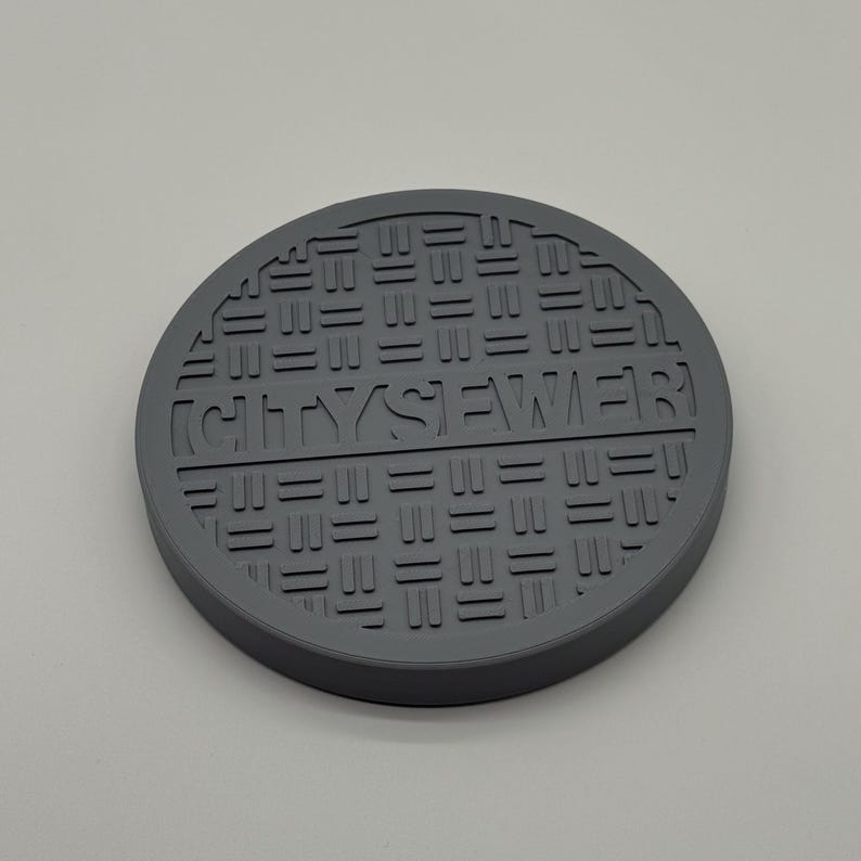 May include: A gray, circular coaster with a manhole cover design. The text "CITY SEWER" is embossed in the center. The coaster has a textured surface and a raised edge.