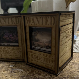 Op de afbeelding: Twee houten dozen met een gezocht-posterontwerp. De posters tonen afbeeldingen van moeraskaarten uit het Magic: The Gathering verzamelkaartspel. De tekst op de posters luidt "WANTED DEAD OR ALIVE" en bevat de namen "PORTGAS-D-ACE" en "MONKEY-D-LUFFY" met beloningsbedragen.