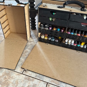 Op de afbeelding: Een houten opbergkoffer met een scharnierend deksel en een handvat. De koffer heeft meerdere laden en vakken om kleine voorwerpen in op te bergen. De koffer is open en de laden zijn gevuld met kleine flesjes verf.