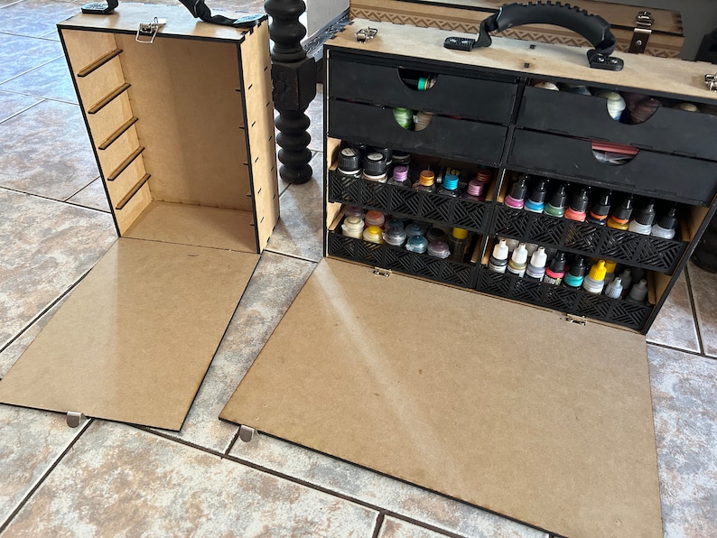 Op de afbeelding: Een houten opbergkoffer met een scharnierend deksel en een handvat. De koffer heeft meerdere laden en vakken om kleine voorwerpen in op te bergen. De koffer is open en de laden zijn gevuld met kleine flesjes verf.