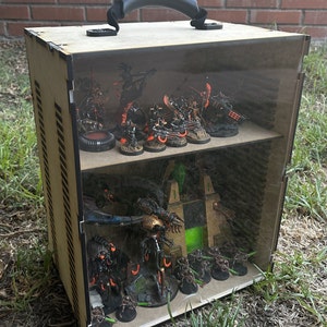 May include: A wooden display case with a clear acrylic lid and a black handle. The case is filled with miniature figures, including a large, multi-legged robot and several smaller figures. The figures are painted in various shades of black, green, and orange.