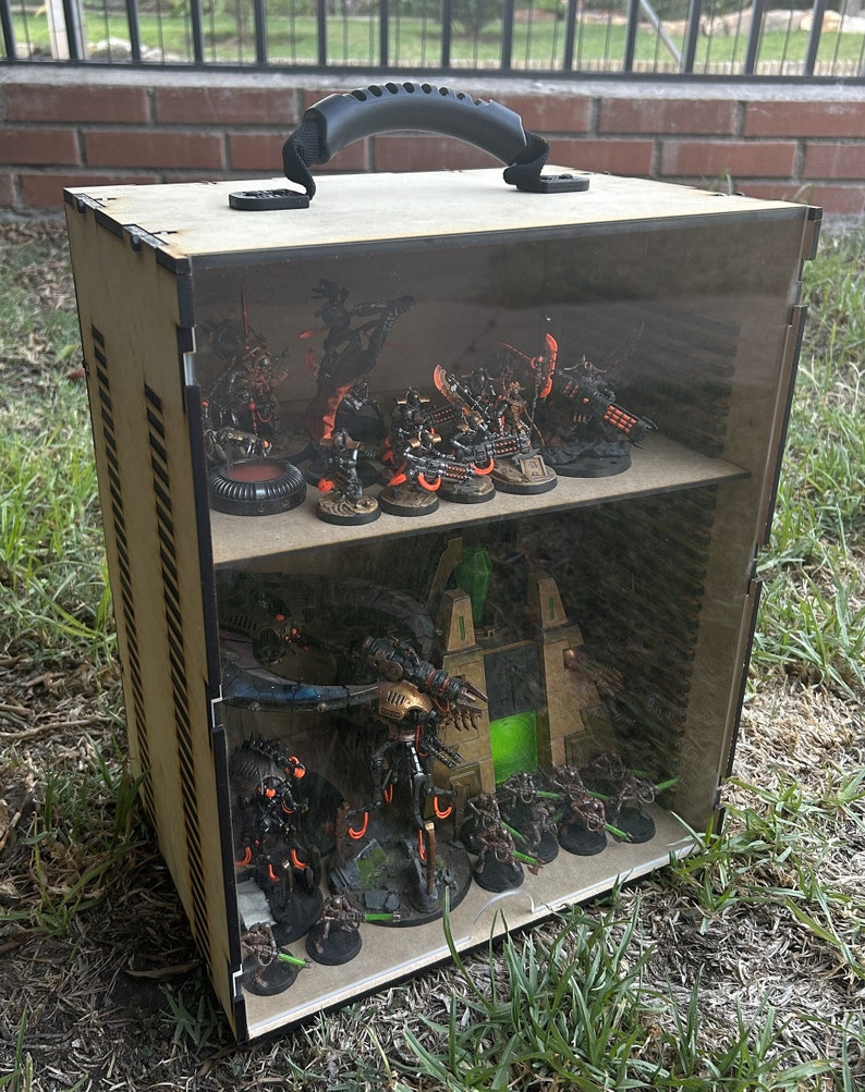 May include: A wooden display case with a clear acrylic lid and a black handle. The case is filled with miniature figures, including a large, multi-legged robot and several smaller figures. The figures are painted in various shades of black, green, and orange.