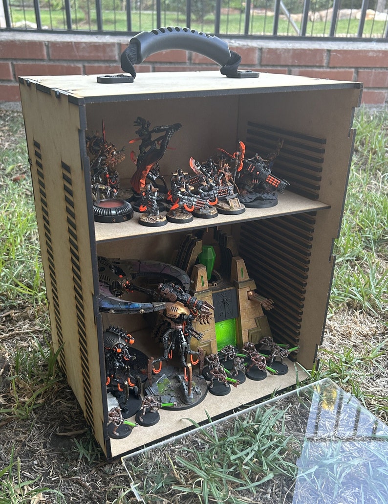 May include: A wooden box with a handle, containing a collection of miniature figures painted in black, orange, and green. The figures are arranged on a shelf inside the box, with a clear plastic lid on top.