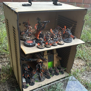 May include: A wooden box with a handle, containing miniature figures of robots and vehicles. The box has two shelves and is painted brown with black accents. The figures are painted in various shades of black, orange, and green.