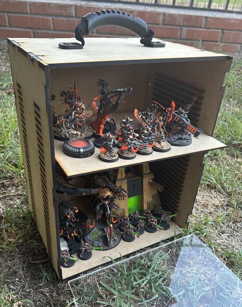 May include: A wooden box with a handle, containing miniature figures of robots and vehicles. The box has two shelves and is painted brown with black accents. The figures are painted in various shades of black, orange, and green.