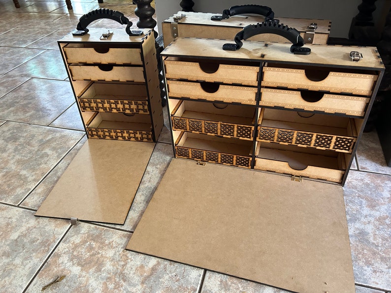 Op de afbeelding: Twee houten opbergdozen met meerdere laden en een scharnierend deksel. De dozen hebben een bruine afwerking en een zwarte handgreep. De dozen zijn open en de laden zijn zichtbaar. De dozen staan op een betegelde vloer.