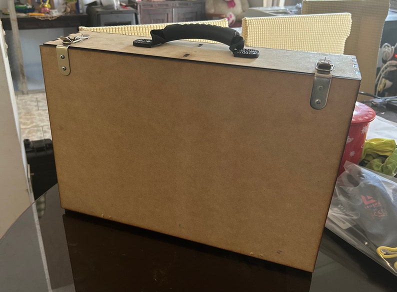 Op de afbeelding: Een bruine houten kist met een zwarte handgreep en zilveren sluitingen. De kist is gesloten en staat op een donker oppervlak.