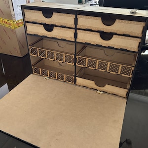 Op de afbeelding: Een houten doos met een scharnierend deksel en meerdere vakken. De doos heeft een zwarte rand en ingewikkelde ontwerpen aan de voorkant. De doos is open en de vakken zijn leeg.