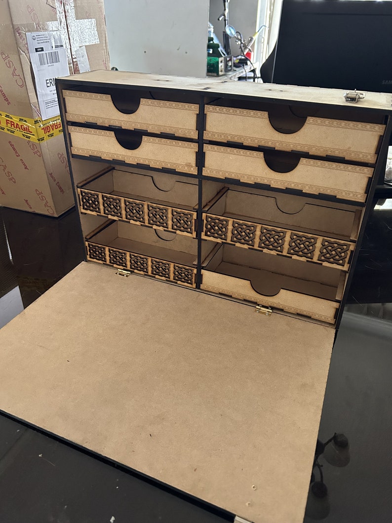 Op de afbeelding: Een houten doos met een scharnierend deksel en meerdere vakken. De doos heeft een zwarte rand en ingewikkelde ontwerpen aan de voorkant. De doos is open en de vakken zijn leeg.