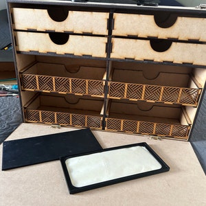 Op de afbeelding: Een houten opbergdoos met meerdere laden en vakken. De doos is open en de laden zijn leeg. De doos heeft een zwarte rand en een zwarte handgreep.