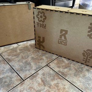 Op de afbeelding: Twee houten dozen met verschillende ontwerpen. De grotere doos heeft een zwarte rand en heeft een geometrisch patroon, de letters "THUY", de letter "R" met "UCS" eronder en een gestileerd symbool. De kleinere doos is simpel.