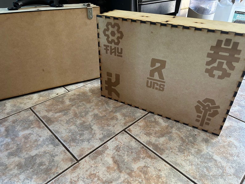 Op de afbeelding: Twee houten dozen met verschillende ontwerpen. De grotere doos heeft een zwarte rand en heeft een geometrisch patroon, de letters "THUY", de letter "R" met "UCS" eronder en een gestileerd symbool. De kleinere doos is simpel.