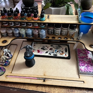 Może przedstawiać: Drewniana stacja do malowania miniaturek z wieloma półkami do przechowywania butelek z farbą. Stacja ma powierzchnię roboczą z paletą do farb, małym kubkiem i pędzlem. Na powierzchni roboczej znajdują się również figurki.