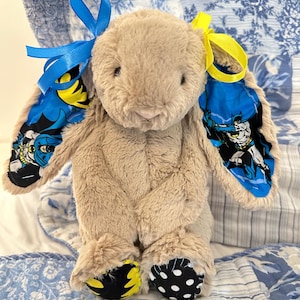 Op de afbeelding: Een pluche konijnenspeelgoed met bruin haar, blauwe en gele Batman-accenten op de oren en zwart-witte stippen en gele accenten op de voeten. Het konijn heeft blauwe en gele linten aan zijn oren.