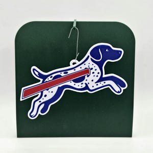 May include: A blue and white dog ornament with a red stripe, jumping across a dark green background. The dog has blue accents and white spots. A silver hook is attached for hanging. The ornament is a decorative item.