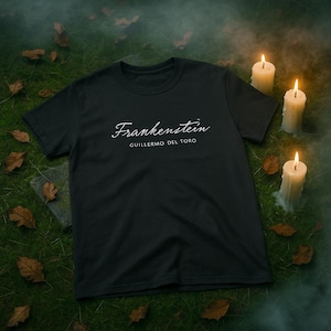 May include: Black t-shirt featuring the word "Frankenstein" in a cursive font, with "Guillermo del Toro" below it. The shirt is displayed on a grassy surface with fallen leaves and lit candles, creating a spooky aesthetic.