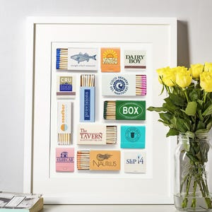 Könnte beinhalten: Ein gerahmter Kunstdruck mit einer Sammlung von Streichholzschachteln mit verschiedenen Logos und Designs, darunter Straight Wharf Restaurant, Dairy Boy, Cisco Brewers, The Tavern, Nautilus und Slip 14, angeordnet in einem Raster auf weißem Grund.