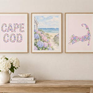 May include: Three framed wall art prints featuring a light pink background. The left print displays the text CAPE COD filled with blue and pink hydrangeas. The centre print shows a coastal beach scene with hydrangeas. The right print shows a map of Cape Cod.