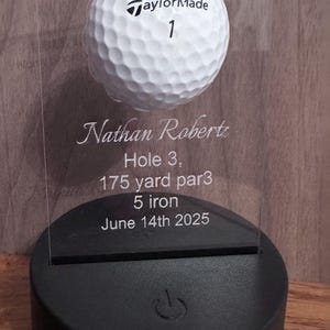 May include: A clear acrylic golf award featuring a white golf ball and the text "HOLE IN ONE". The award includes the name "Nathan Roberts", hole details, and the date June 14th, 2025. It is mounted on a black base.