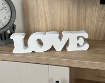 May include: A white, three-dimensional wooden sign spells out the word "LOVE" in capital letters. The letters are thick and stand upright on a wooden surface. A decorative clock is visible in the background.