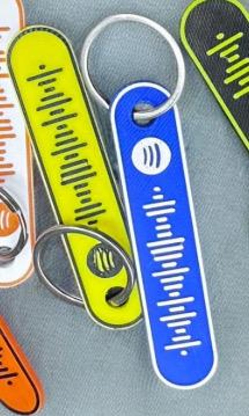 K&ouml;nnte beinhalten: Schl&uuml;sselanh&auml;nger in verschiedenen Farben, darunter Gelb und Blau, jeweils mit einem einzigartigen Audio-Wellenform-Design und einem Metall-Schl&uuml;sselring. Der blaue Schl&uuml;sselanh&auml;nger zeigt ein wei&szlig;es Spotify-Logo. Die Schl&uuml;sselanh&auml;nger sind rechteckig mit abgerundeten Kanten.