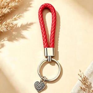 May include: A red braided cord keyring featuring a silver-toned metal connector and a circular split ring. Attached to the ring is a small, heart-shaped charm with an intricate, embossed filigree pattern in a dark, oxidised silver finish.