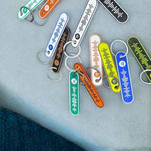 K&ouml;nnte beinhalten: Eine Sammlung farbenfroher Spotify-Code-Schl&uuml;sselanh&auml;nger in verschiedenen Farben, darunter Gr&uuml;n, Orange, Schwarz, Wei&szlig;, Blau und Gelb. Jeder Schl&uuml;sselanh&auml;nger hat ein einzigartiges Spotify-Code-Design und einen silbernen Schl&uuml;sselring. Die Schl&uuml;sselanh&auml;nger liegen auf einer hellgrauen Oberfl&auml;che.