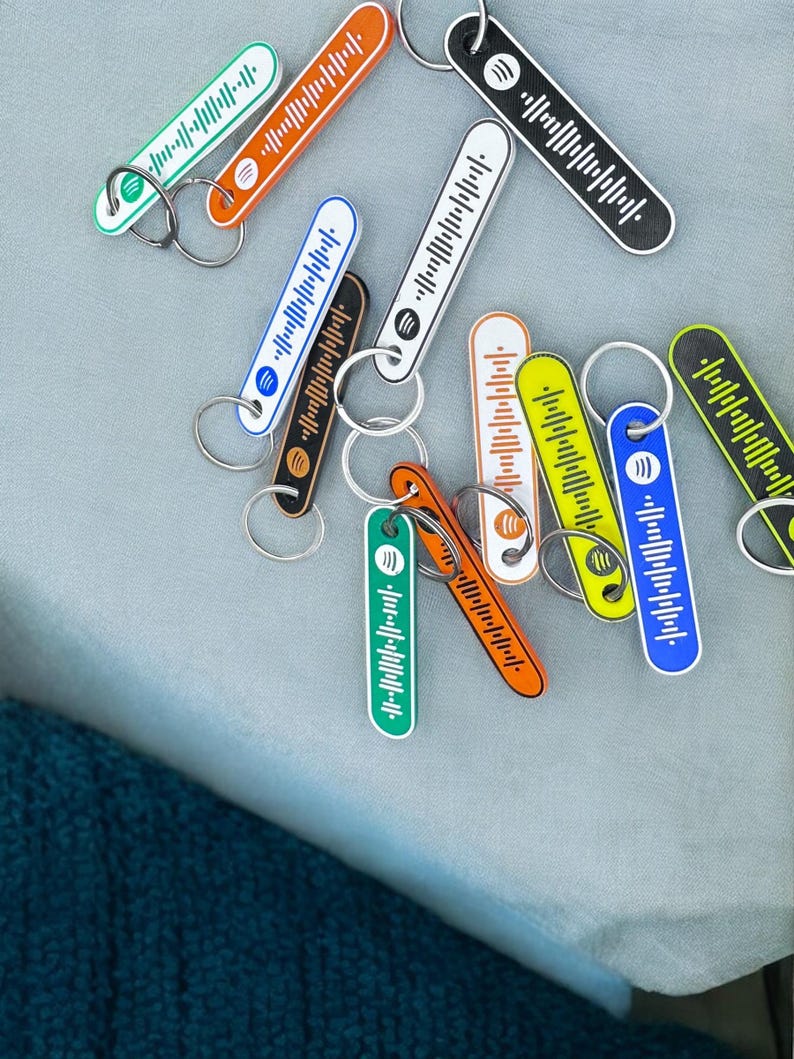 K&ouml;nnte beinhalten: Eine Sammlung farbenfroher Spotify-Code-Schl&uuml;sselanh&auml;nger in verschiedenen Farben, darunter Gr&uuml;n, Orange, Schwarz, Wei&szlig;, Blau und Gelb. Jeder Schl&uuml;sselanh&auml;nger hat ein einzigartiges Spotify-Code-Design und einen silbernen Schl&uuml;sselring. Die Schl&uuml;sselanh&auml;nger liegen auf einer hellgrauen Oberfl&auml;che.