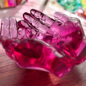 Op de afbeelding: Een roze glazen kom in de vorm van twee samen gevouwen handen. De kom is gemaakt van helder glas met een roze tint.