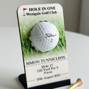 Op de afbeelding: Een golfbaldisplay met een Titleist golfbal. De display toont "HOLE IN ONE" en "Wesigale Golf Club". Details zijn: Hole 17, 110.9 meter Par 3, 9 ijzer en de datum. Een houten golf-tee is ook inbegrepen.