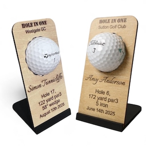 May include: Two wooden golf ball displays. Each display holds a golf ball and features engraved text. One says "Hole in One" and the other says "TaylorMade 1". The other says "Titleist 3". The displays have black bases.