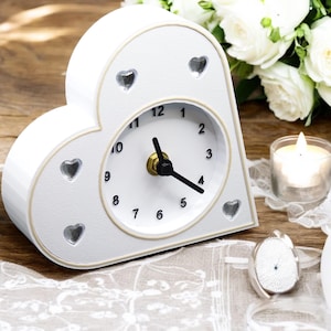 Peut inclure: Horloge en forme de cœur blanche avec un cadran blanc et des aiguilles noires. L'horloge a une bordure dorée et un motif de cœur décoratif sur le devant. L'horloge est posée sur une nappe en dentelle blanche.