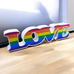 May include: White LOVE sign with rainbow stripes. The sign is made of plastic and is 3D.