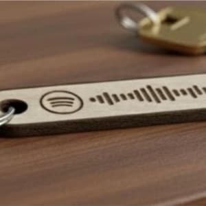 May include: Wooden Spotify keyring with a silver key ring. The keyring features the Spotify logo and a carved sound wave design. A gold key is in the background. The keyring is made of light-coloured wood with a dark brown edge.