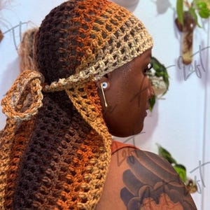 以下が含まれることがあります： 茶色、オレンジ、ベージュのクロシェ編みのヘアースカーフで、ゆったりとした流れるようなデザインです。スカーフは人の頭に巻かれ、背中に垂れ下がっています。