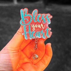 May include: A teal and pink glitter badge holder with a retractable badge reel. The badge holder has the text "Bless your heart" in a white script font.