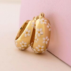 May include: Gold-colored hoop earrings with a white daisy pattern. The earrings have a rounded shape and a glossy finish. The daisies are evenly spaced across the surface of the earrings. The earrings are displayed against a pink background.