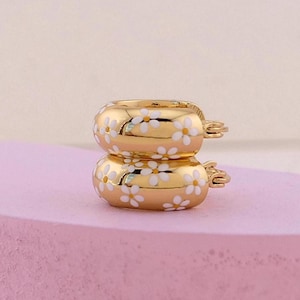 May include: Two gold-colored hoop earrings with a white daisy pattern. The earrings are stacked on top of each other, showcasing their design. The background is a soft pink and beige.