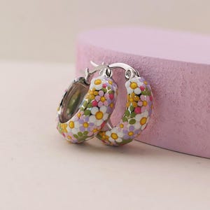 May include: A pair of silver hoop earrings featuring a colourful enamel floral pattern. The design includes small white, yellow, pink, and light purple daisy-like flowers covering the outer surface of the thick, rounded metal hoops.