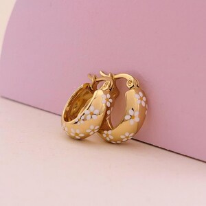 May include: A pair of gold-colored hoop earrings with a white daisy floral pattern. The earrings are set against a pink background. The earrings are a jewelry item.