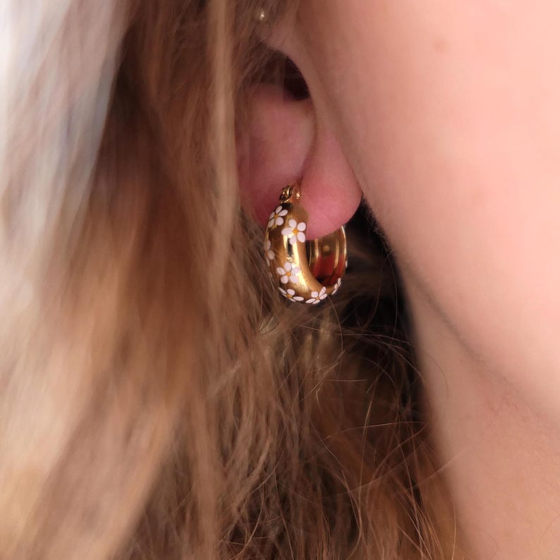 May include: Gold-colored hoop earring with a white floral pattern. The earring is a thick, rounded shape, and the white flowers are evenly spaced around the outer surface. The earring is worn on the ear.