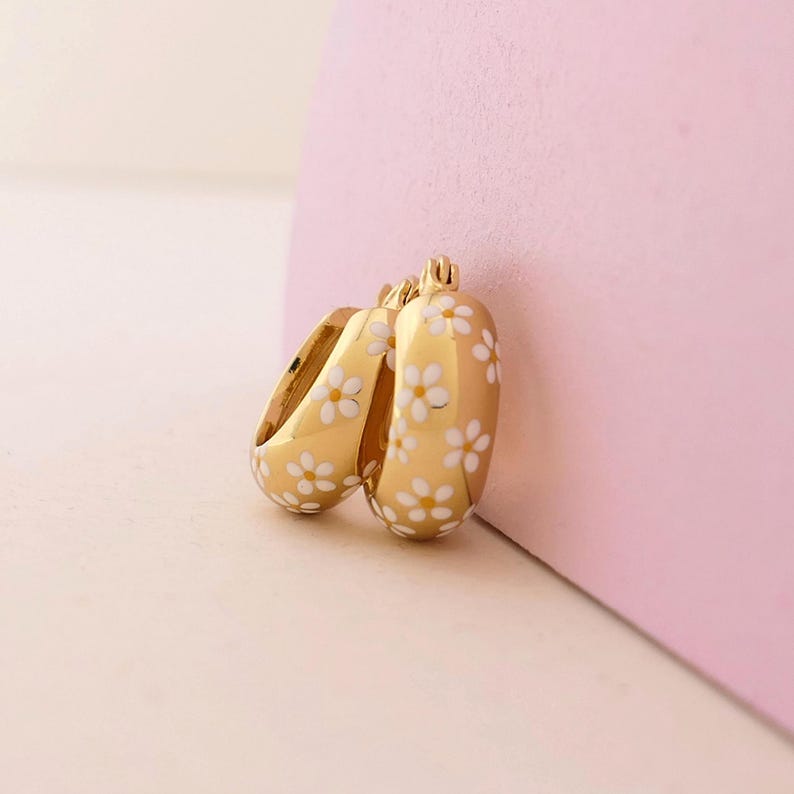 May include: Gold-colored hoop earrings with a white daisy pattern. The earrings have a rounded shape and a glossy finish. The daisies are evenly spaced across the surface of the earrings. The earrings are displayed against a pink background.