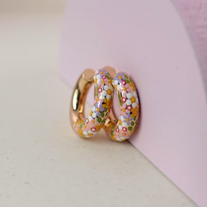 Puede incluir: Un par de pendientes de aro dorados con un dise&ntilde;o floral. Los pendientes est&aacute;n cubiertos de peque&ntilde;as flores coloridas en tonos de rosa, morado, amarillo y verde.
