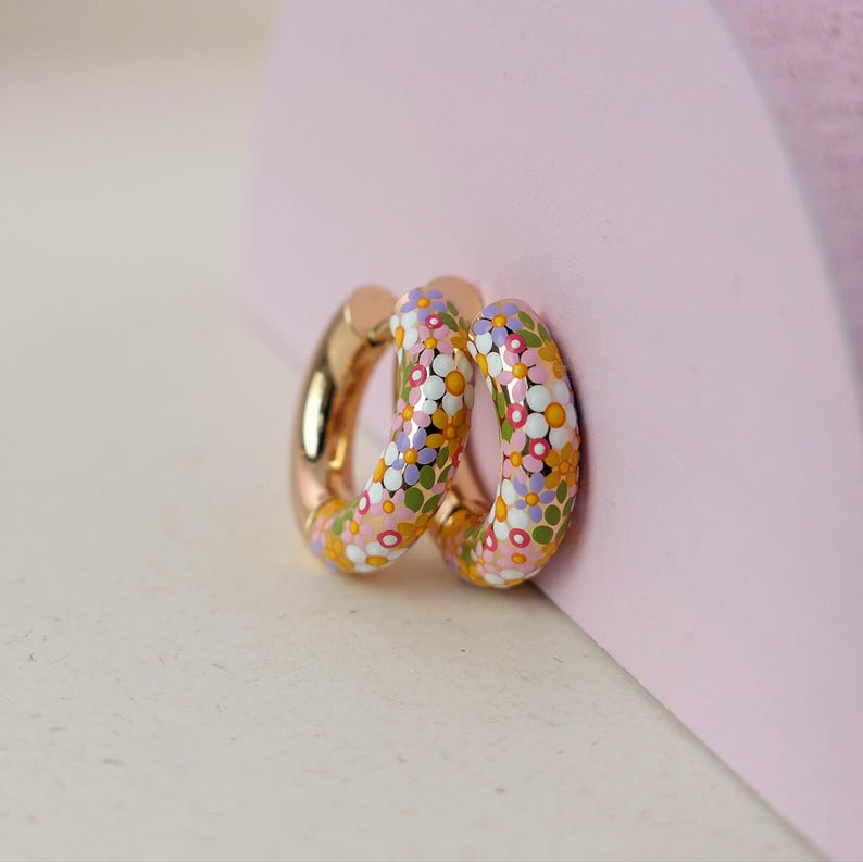 Puede incluir: Un par de pendientes de aro dorados con un dise&ntilde;o floral. Los pendientes est&aacute;n cubiertos de peque&ntilde;as flores coloridas en tonos de rosa, morado, amarillo y verde.