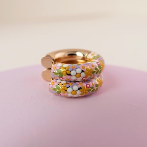 Puede incluir: Un par de pendientes de aro dorados con un dise&ntilde;o floral. Los pendientes est&aacute;n cubiertos con un patr&oacute;n colorido de flores rosas, amarillas, blancas y verdes.
